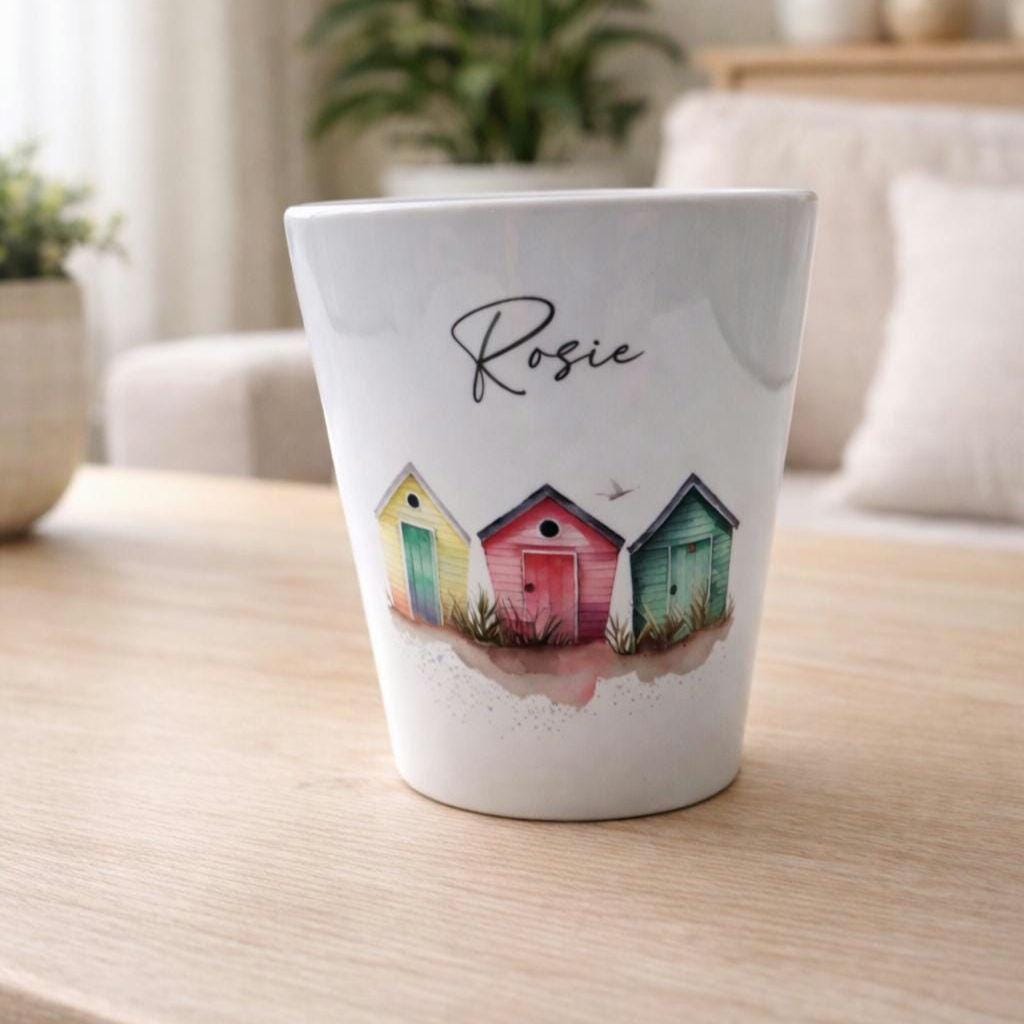 White plant pot with colourful beach huts design and 'Rosie' text on a wooden surface.